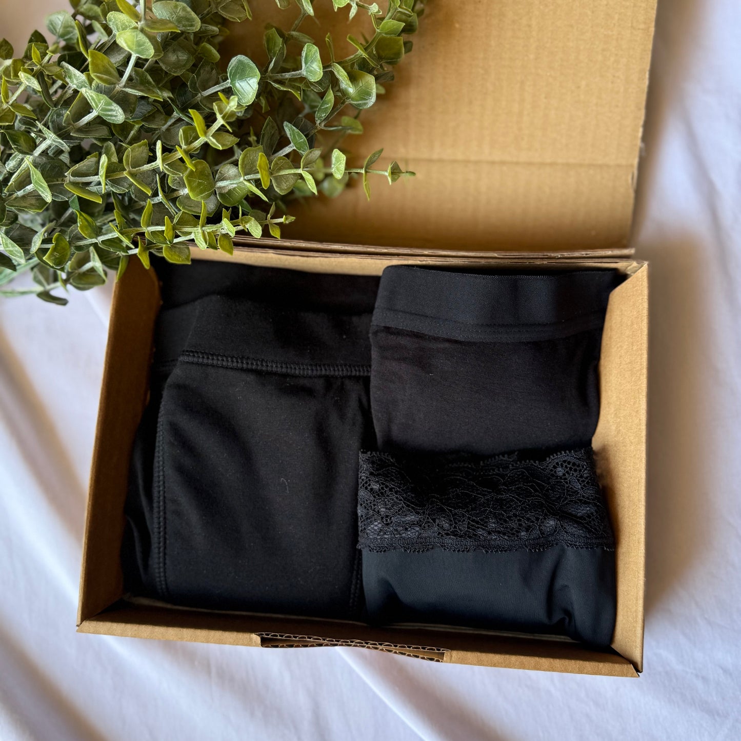 Coffret découverte féminité dans sa boite, présenté sur un drap blanc et décoré de branches vertes, vu de dessus.