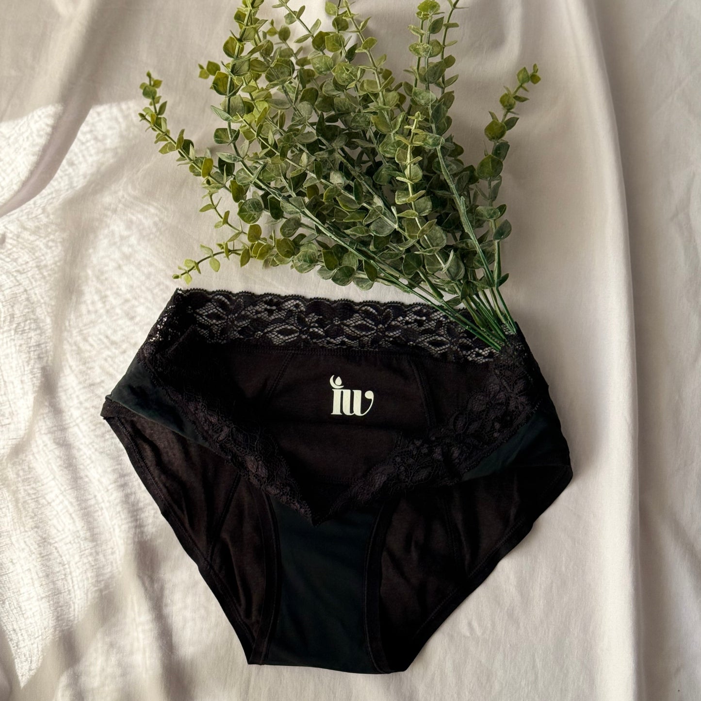 Culotte menstruelle dentelle présentée sur un drap blanc, décorée de branches sous une lumière claire.