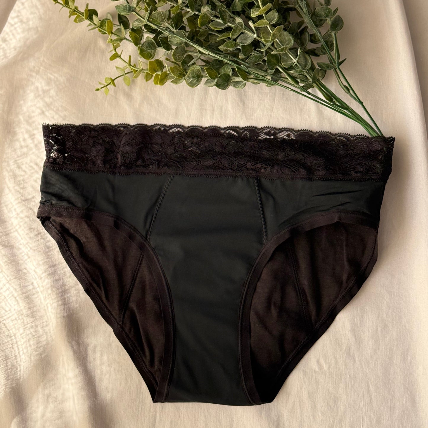 Culotte menstruelle dentelle présentée sur un drap blanc, décorée de branches, sous une lumière claire.