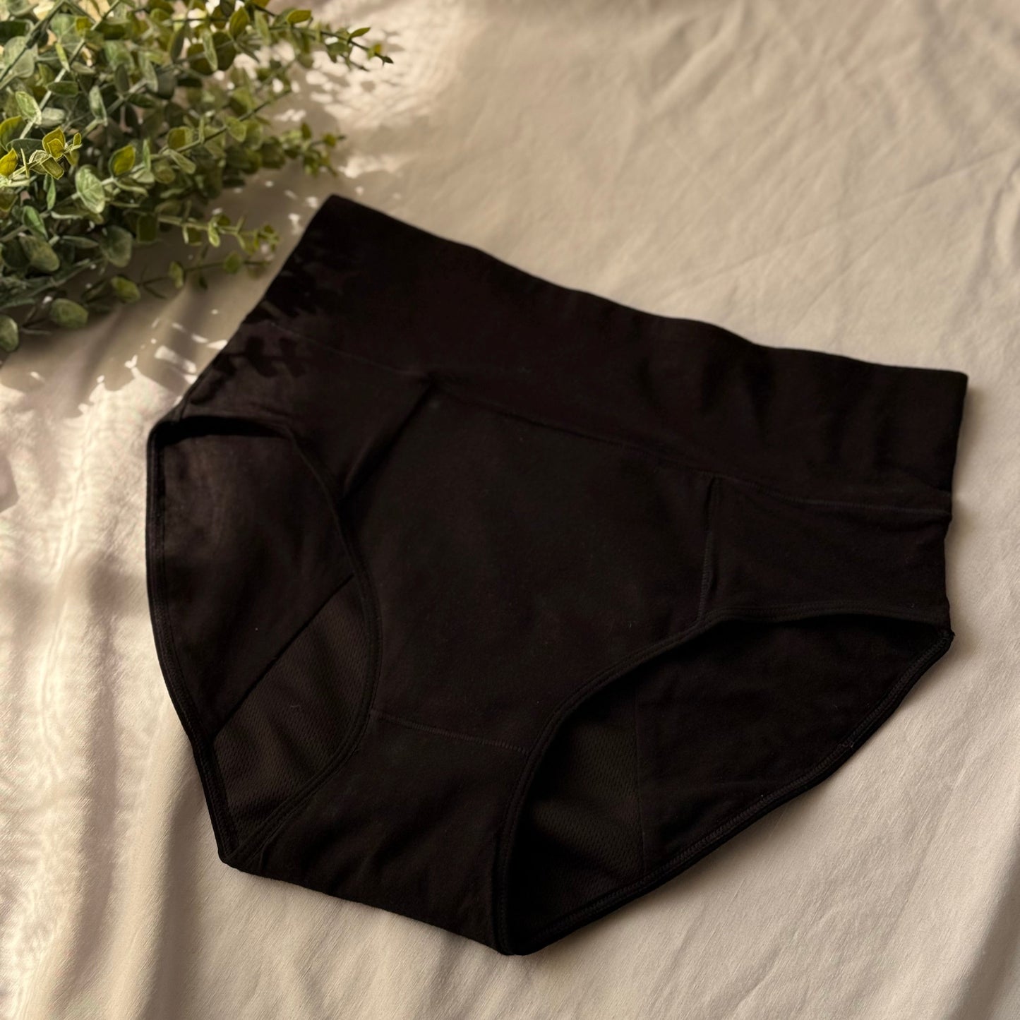 Culotte menstruelle taille haute, prise de face, présentée sur un drap blanc et décorée d'une branche.