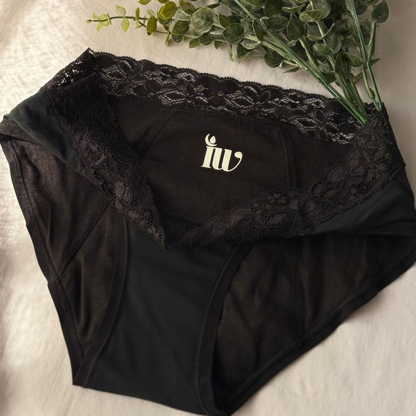 Culotte menstruelle dentelle présentée sur un drap blanc, décorée de branche, avec l'encolure pliée laissant apparaitre le logo.