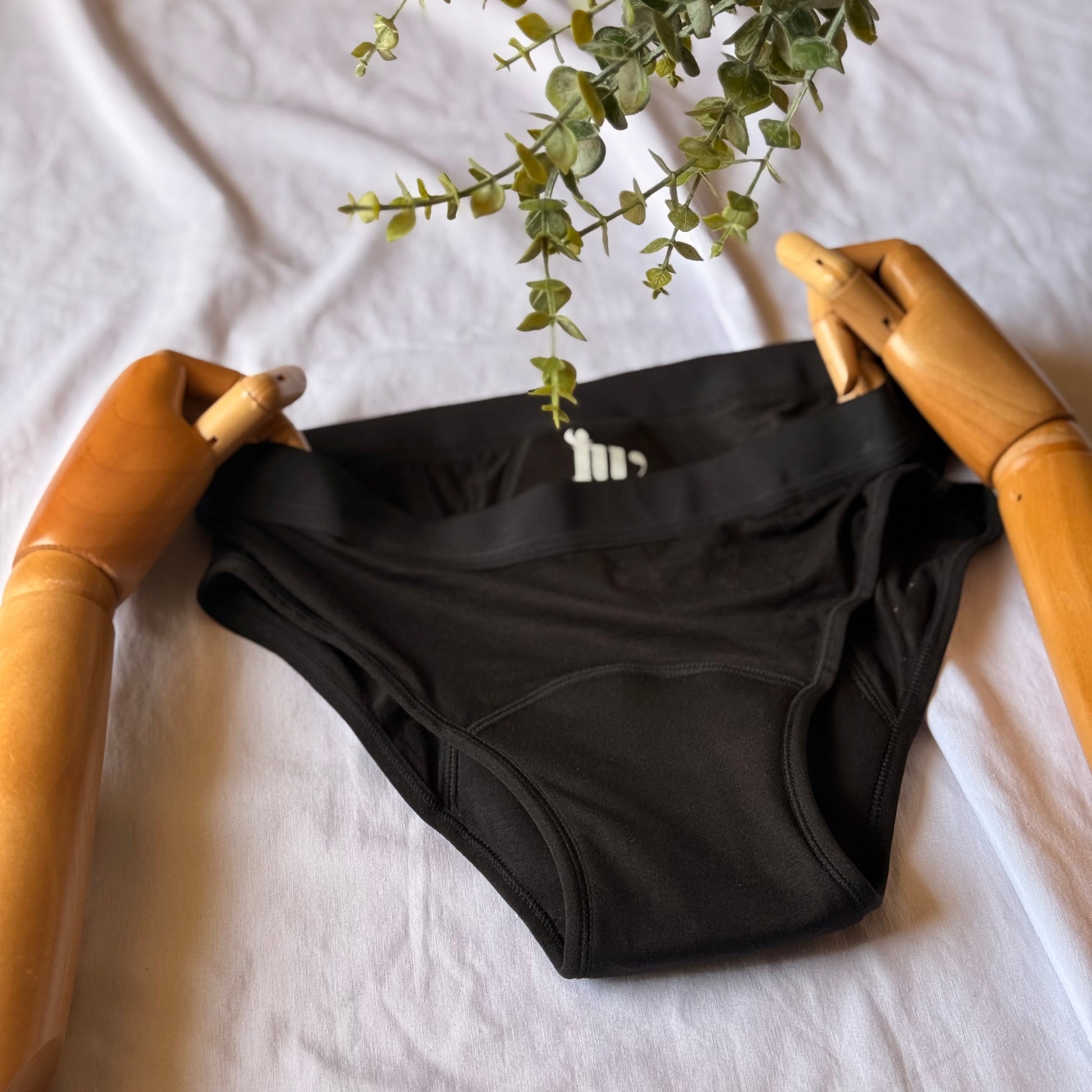 Culotte menstruelle standard présentée par un mannequin articulé en bois, sur un drap blanc, mettant en avant le gousset de la culotte, avec des feuilles visibles sur le haut de l'image.