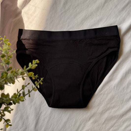 Culotte menstruelle standard présentée sur un drap blanc, avec de branches feuillues visibles sur le côté de l'image, avec une lumière visible au coin.