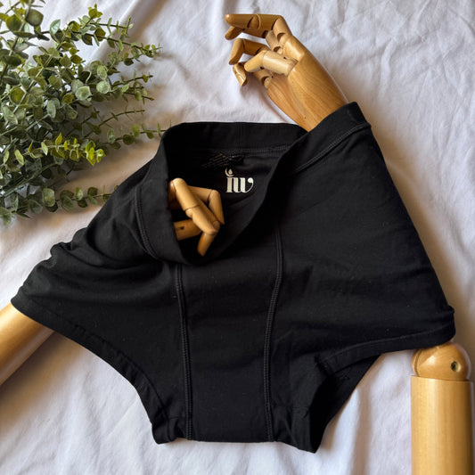 Culotte menstruelle modèle nuit, présentée sur un mannequin en bois articulé, posée sur un drap blanc avec une branche de feuillage en décoration.