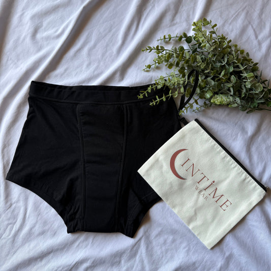 Pochette intime wear beige présentée sur un drap blanc, accompagnée d'une culotte nuit et décorée de branches vertes.