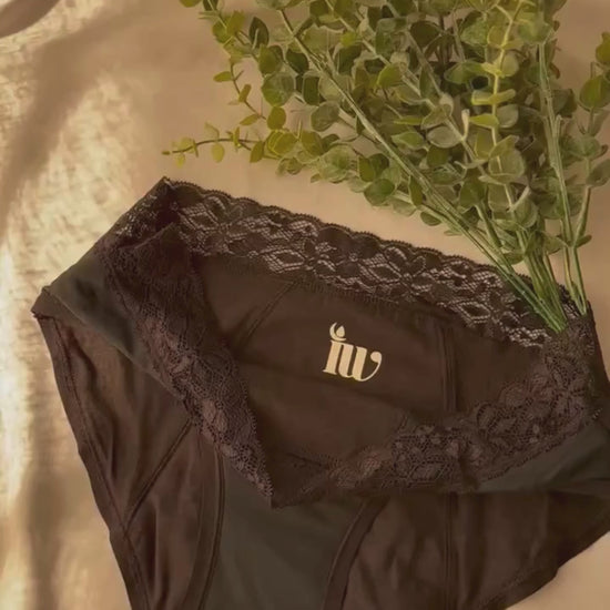 Culotte menstruelle dentelle présentée sur un drap blanc et décorée de branches, dans une lumière tamisée.