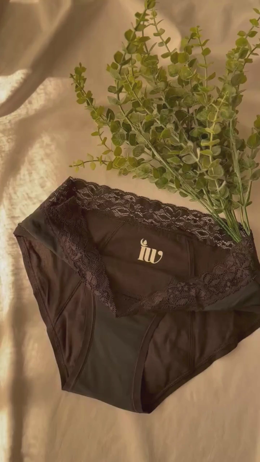 Culotte menstruelle dentelle présentée sur un drap blanc et décorée de branches, dans une lumière tamisée.